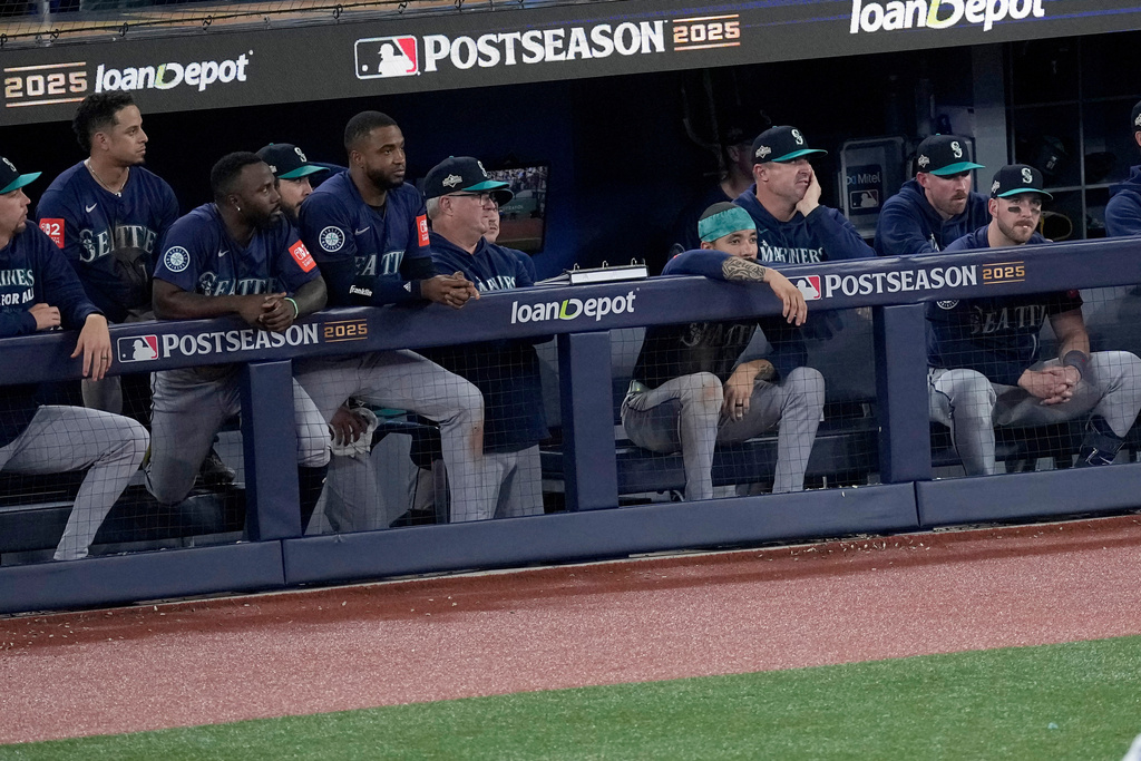 Seattle Mariners players in the dugout during a postseason game, leaning on the railing and watching the field, with Postseason 2025 and loanDepot signage visible.