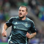 Soccer player in a dark jersey running on the field, Providence sponsor visible on chest, blurred stadium crowd in background