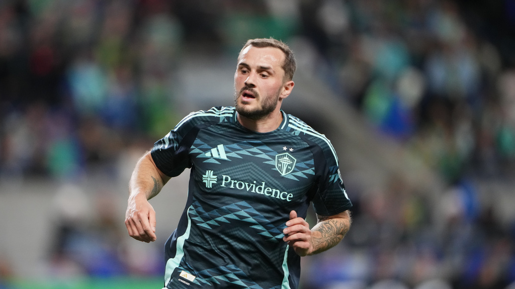 Soccer player in a dark jersey running on the field, Providence sponsor visible on chest, blurred stadium crowd in background