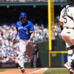 Kansas City Royals runner in blue sprinting toward home plate as the catcher in white gear stands nearby with a packed stadium in the background.