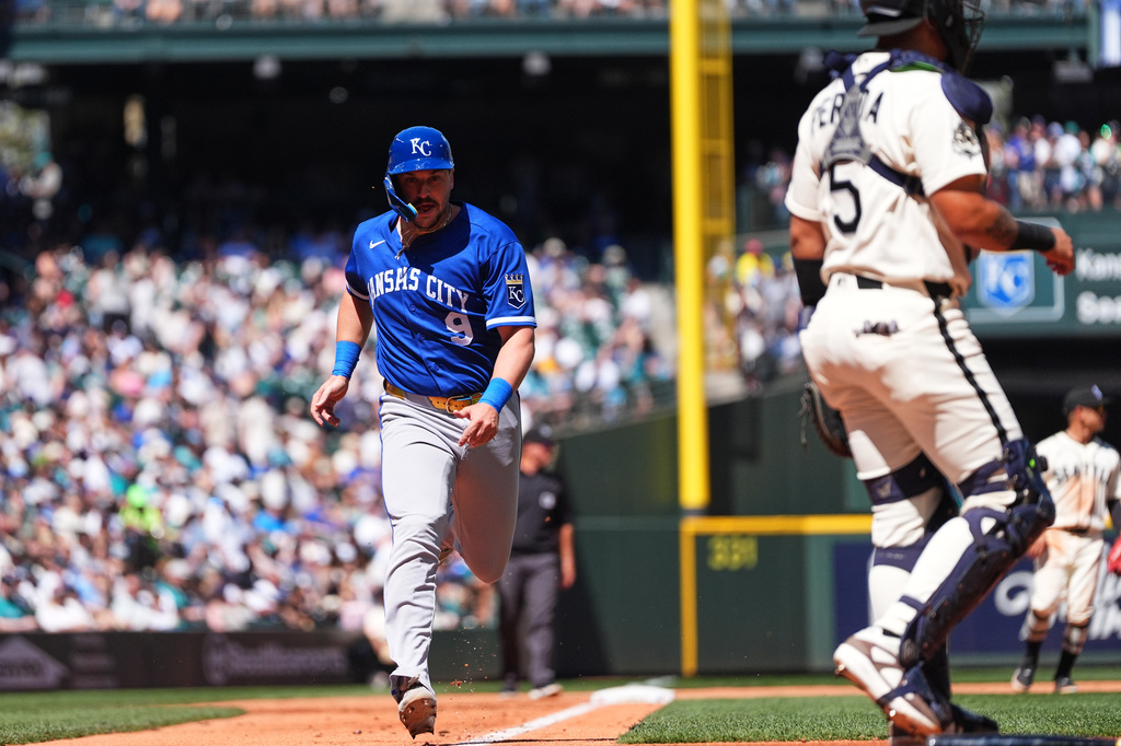 Kansas City Royals runner in blue sprinting toward home plate as the catcher in white gear stands nearby with a packed stadium in the background.