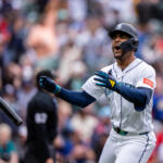 Seattle Mariners batter in white uniform swings as the bat flies off to the left, crowd blurred in the stadium background.