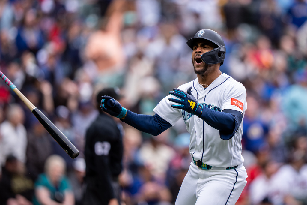 Seattle Mariners batter in white uniform swings as the bat flies off to the left, crowd blurred in the stadium background.