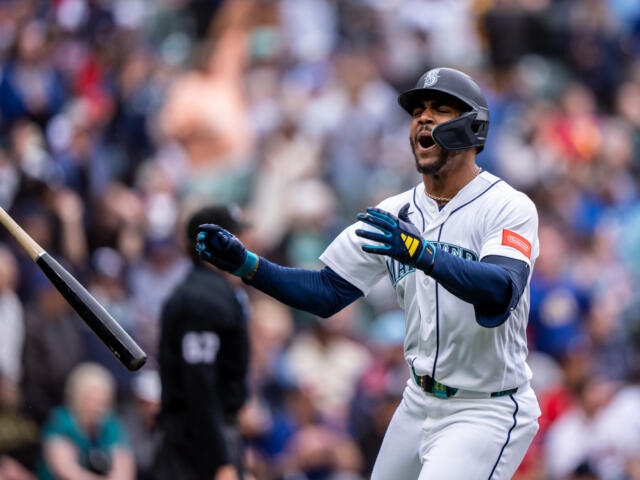 Mariners batter in white uniform celebrates as his bat sails through the air in the foreground and a cheering crowd watches