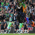 Soccer players in green and dark uniforms celebrate a goal on the field while a packed stadium fans raise their arms in celebration behind them.