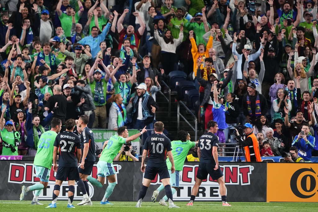 Soccer players in green and dark uniforms celebrate a goal on the field while a packed stadium fans raise their arms in celebration behind them.
