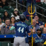 Baseball player Rodríguez #44 in blue uniform stands at the dugout, holding a tall gold trident while a teammate looks on.