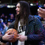 Man with long dreadlocks in a patterned jacket holds a basketball on the court as spectators watch in the background.