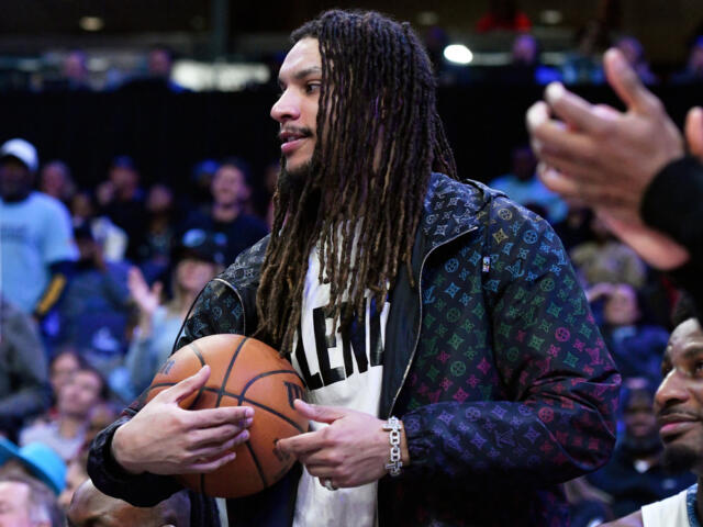 Man with long dreadlocks in a patterned jacket holds a basketball on the court as spectators watch in the background.