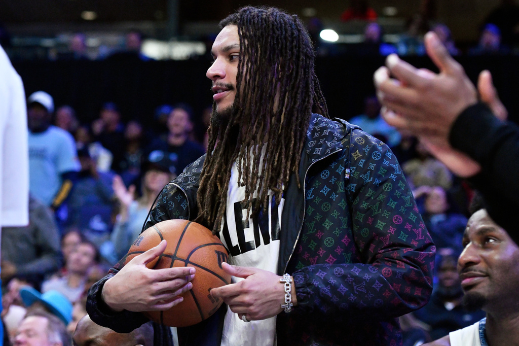 Man with long dreadlocks in a patterned jacket holds a basketball on the court as spectators watch in the background.