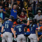 Baseball players in blue jerseys celebrate in the dugout as one raises a gold trident overhead while fans cheer behind them in the stands.
