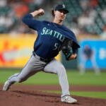 A Seattle Mariners pitcher in a blue uniform winds up on the mound and throws a pitch, with a blurred stadium crowd in the background.