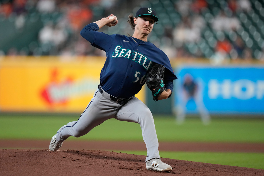 A Seattle Mariners pitcher in a blue uniform winds up on the mound and throws a pitch, with a blurred stadium crowd in the background.