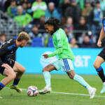 Soccer players contest the ball on a green pitch as a player in a bright green jersey advances with the ball while opponents in dark blue kits close in, with a crowd in the stands behind.