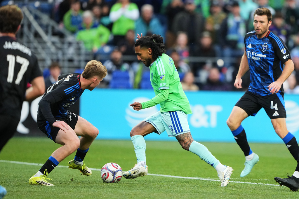 Soccer players contest the ball on a green pitch as a player in a bright green jersey advances with the ball while opponents in dark blue kits close in, with a crowd in the stands behind.