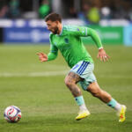 Soccer player in a neon green jersey and light shorts runs toward a ball on a grassy field during a match, tattoos visible on his leg.