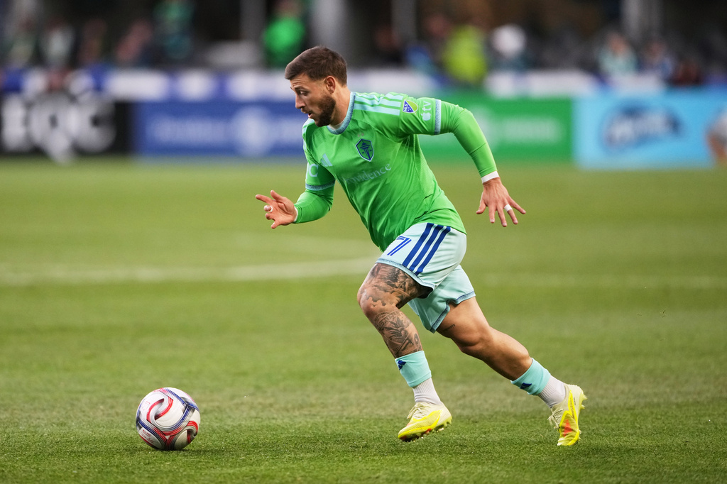 Soccer player in a neon green jersey and light shorts runs toward a ball on a grassy field during a match, tattoos visible on his leg.