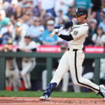 Baseball batter in a white uniform swinging at a pitch, crowd watching from the stands behind him.