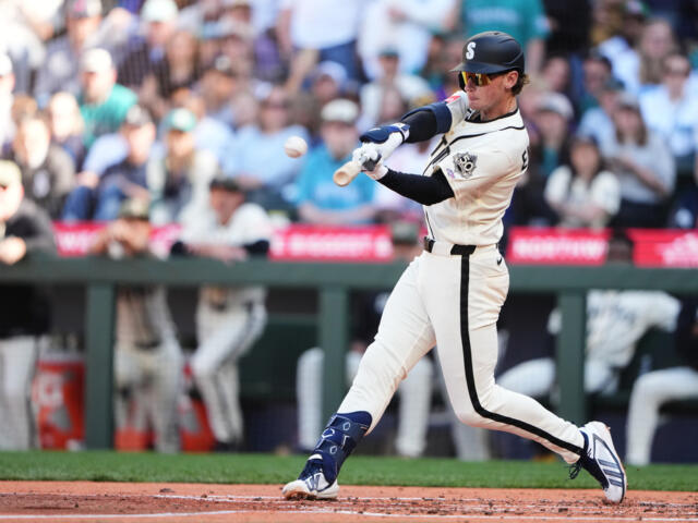 Baseball batter in a white uniform swinging at a pitch, crowd watching from the stands behind him.
