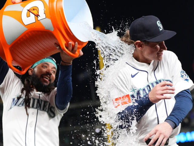 Two Seattle Mariners players celebrate, pouring an orange Gatorade jug over one player's head as water sprays around them in white uniforms.