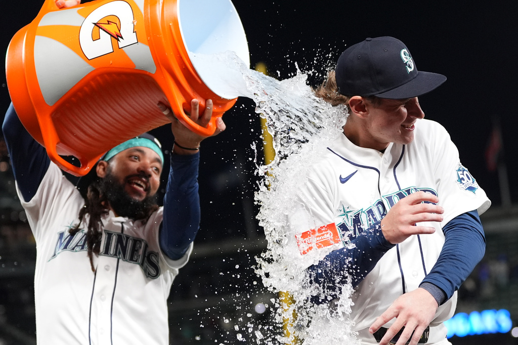Two Seattle Mariners players celebrate, pouring an orange Gatorade jug over one player's head as water sprays around them in white uniforms.