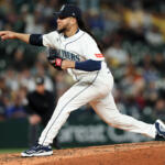 Seattle Mariners pitcher in white uniform mid-pitch on the mound during a game with a blurred crowd in the background.