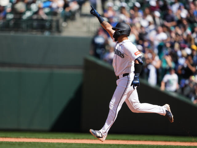 Baseball player in a white uniform runs along the baseline with one arm raised, as fans watch from the stands.