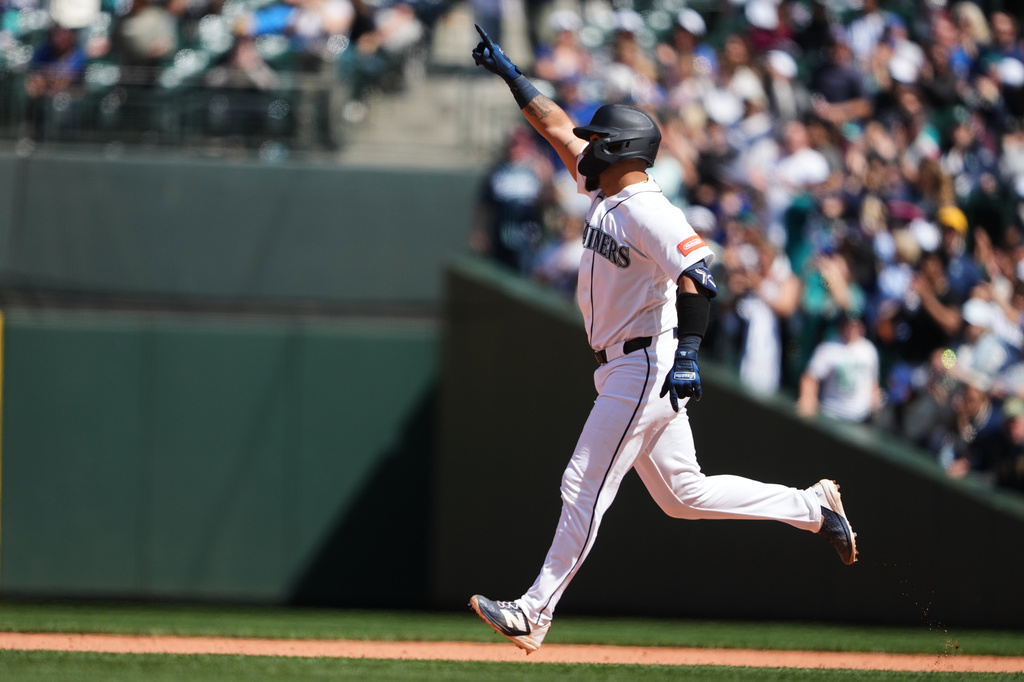 Baseball player in a white uniform runs along the baseline with one arm raised, as fans watch from the stands.