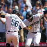 Two Seattle Mariners players in white uniforms high-fiving on the field as fans cheer nearby.