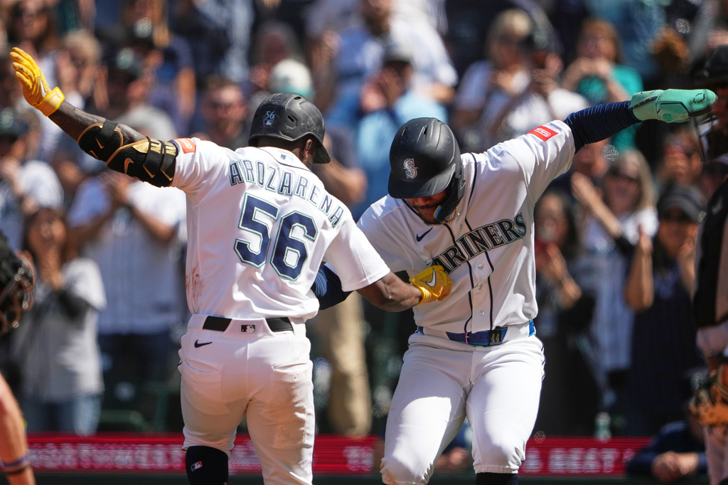 Two Seattle Mariners players in white uniforms high-fiving on the field as fans cheer nearby.