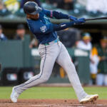Mariners batter in blue swings a baseball bat during a game, mid-swing with a catcher visible on the left field side and players in the dugout in the background.