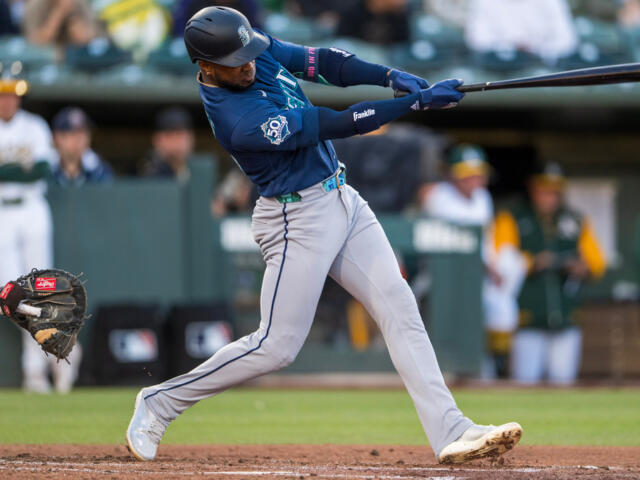 Mariners batter in blue swings a baseball bat during a game, mid-swing with a catcher visible on the left field side and players in the dugout in the background.