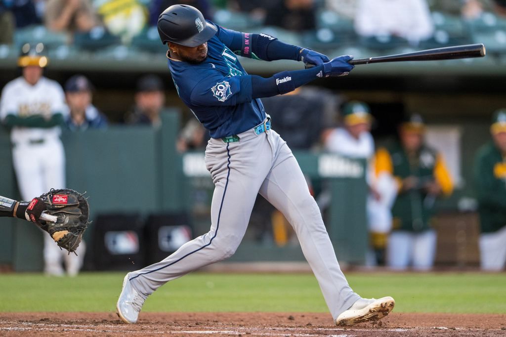 Mariners batter in blue swings a baseball bat during a game, mid-swing with a catcher visible on the left field side and players in the dugout in the background.