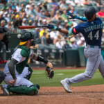 Baseball batter swinging at a pitch as catcher and umpire react behind him with a crowd in the stands.