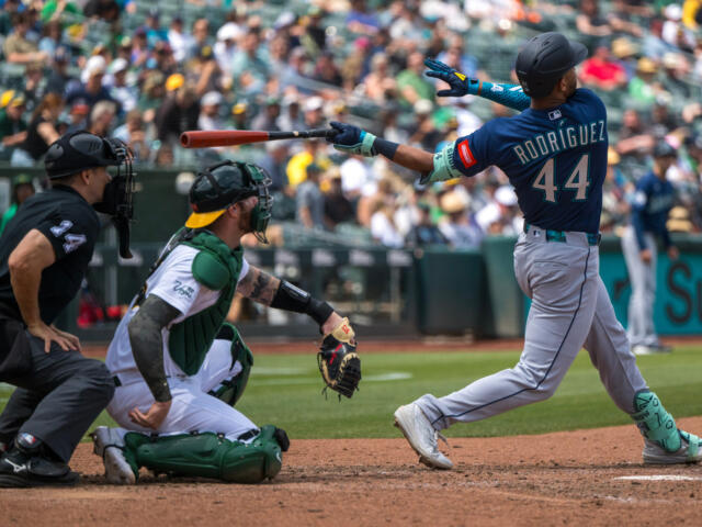Baseball batter swinging at a pitch as catcher and umpire react behind him with a crowd in the stands.