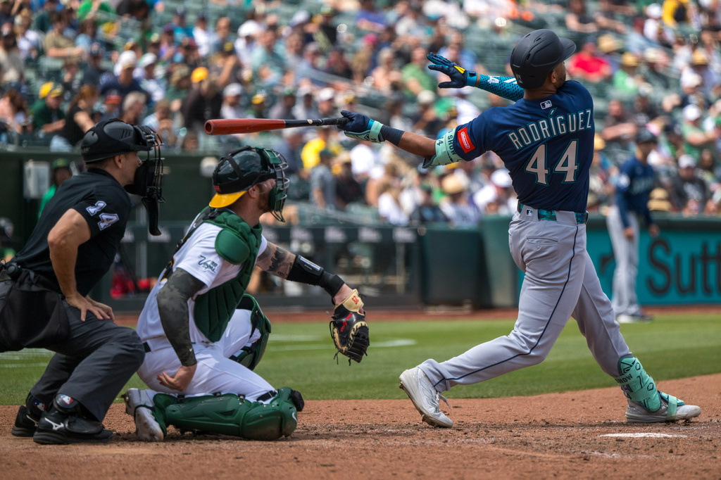 Baseball batter swinging at a pitch as catcher and umpire react behind him with a crowd in the stands.