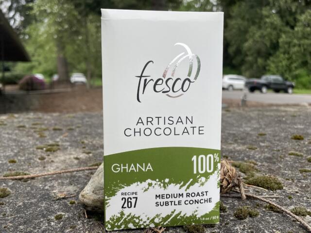 Close-up of a Fresco artisan chocolate box showing Ghana 100% with green accent at the bottom outside on the ground.