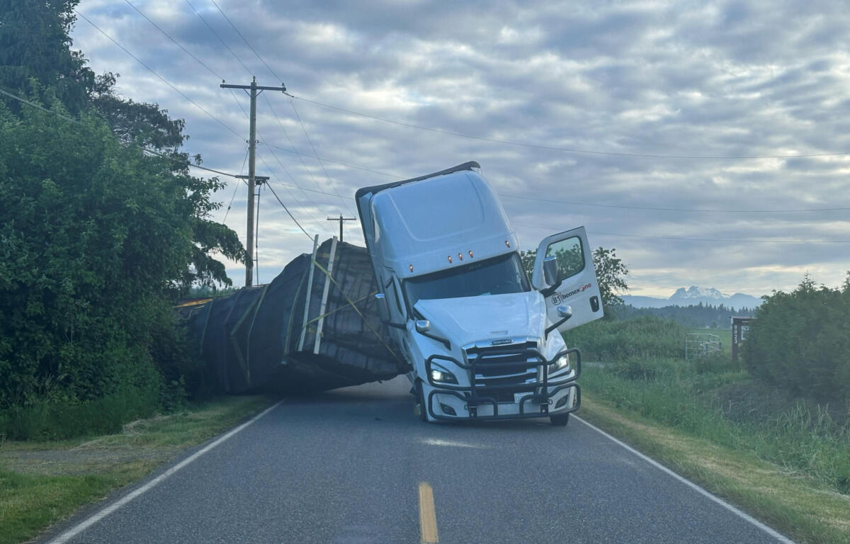 Blue semi-truck leaning on its side across a rural two-lane road with power lines and trees nearby, cloudy sky above.