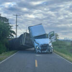 Blue semi-truck leaning on its side across a rural two-lane road with power lines and trees nearby, cloudy sky above.