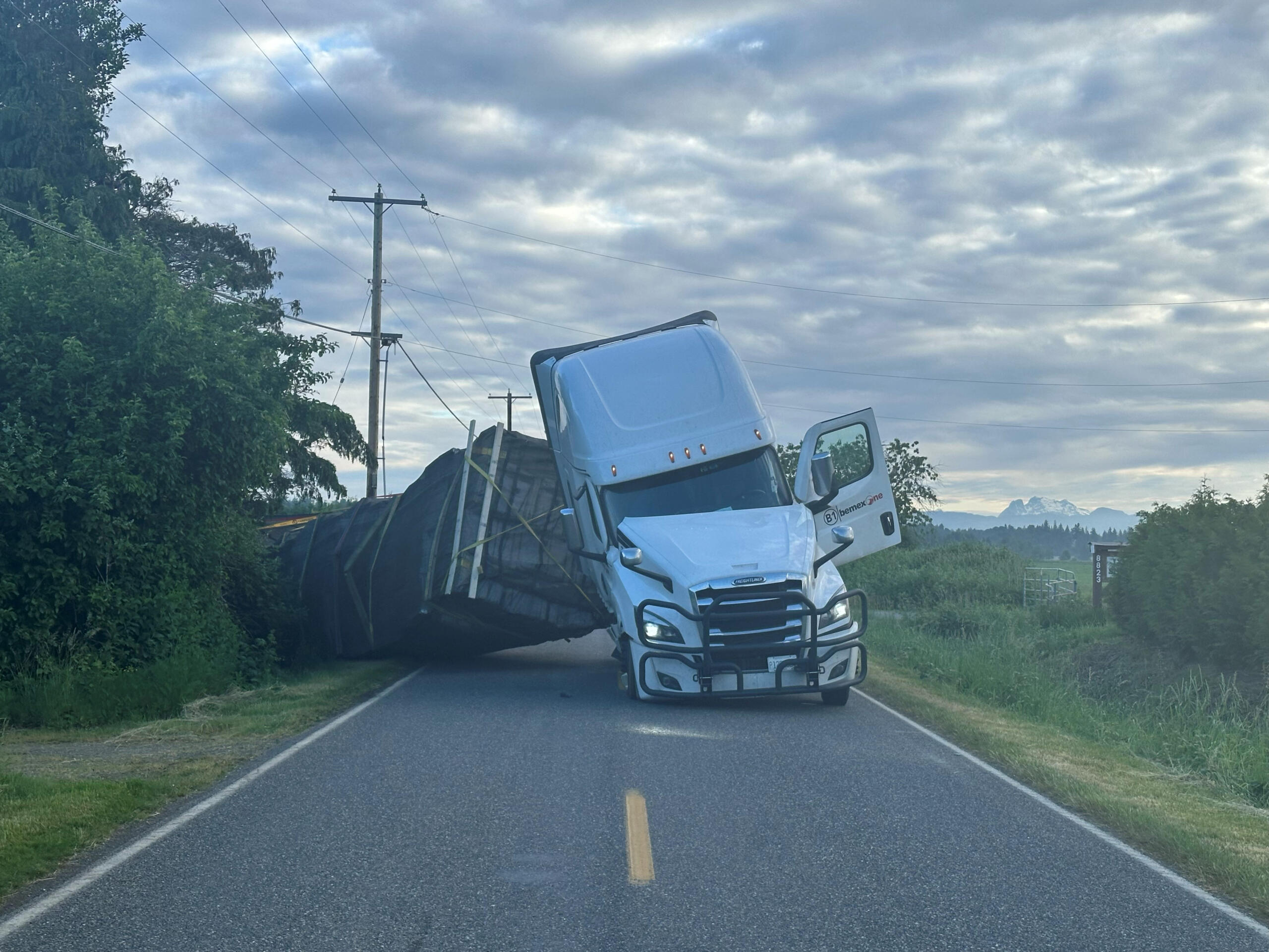 Blue semi-truck leaning on its side across a rural two-lane road with power lines and trees nearby, cloudy sky above.