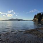 Coastal scene with calm water, a pebble beach, and a forested rocky cliff on the right under a clear blue sky.