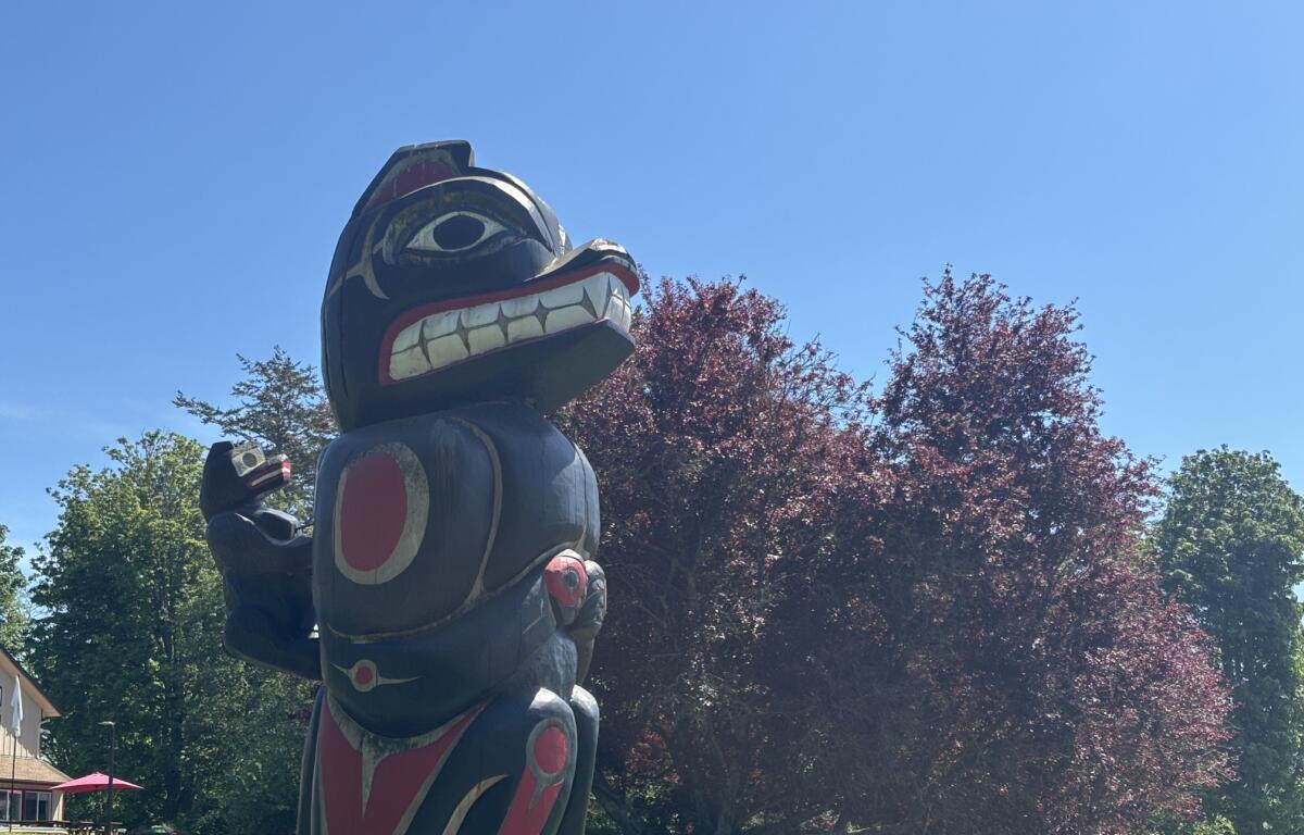 Large wooden totem pole sculpture with bold red, white, and black carvings, outdoors in a sunny park against a blue sky.
