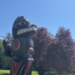 Large wooden totem pole sculpture with bold red, white, and black carvings, outdoors in a sunny park against a blue sky.