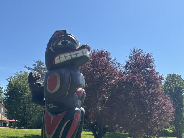 Large wooden totem pole sculpture with bold red, white, and black carvings, outdoors in a sunny park against a blue sky.
