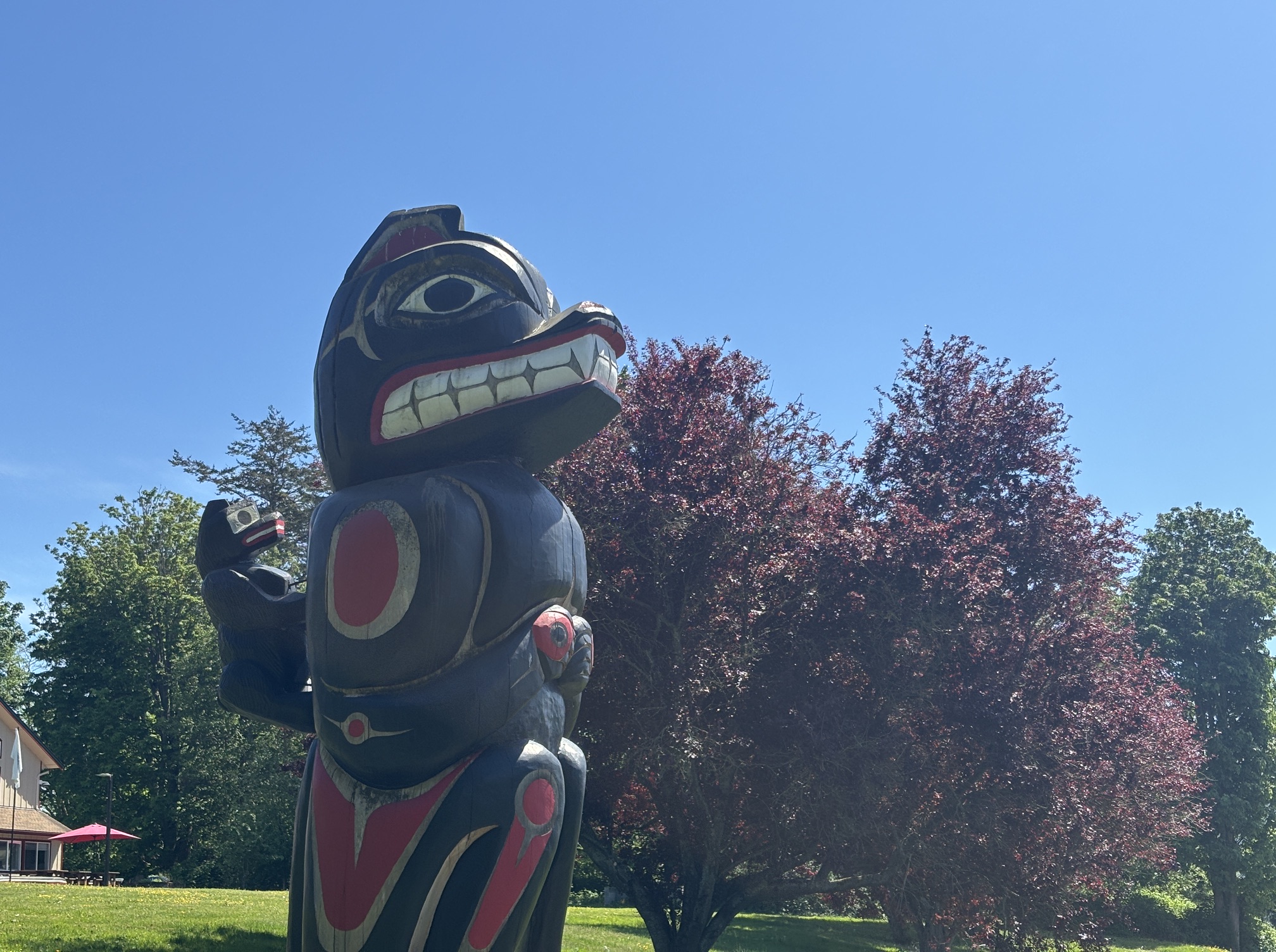Large wooden totem pole sculpture with bold red, white, and black carvings, outdoors in a sunny park against a blue sky.
