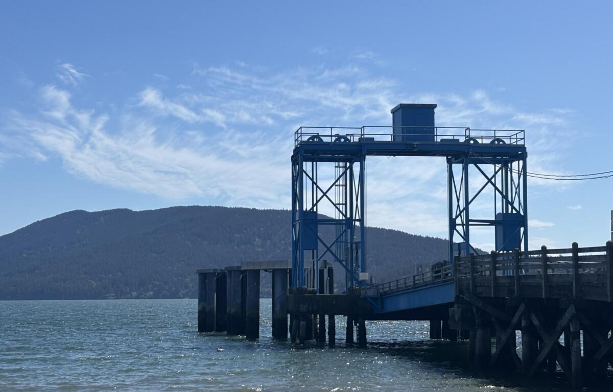 Blue industrial pier structure stretching over the water with a mountainous coastline in the distance and a clear blue sky