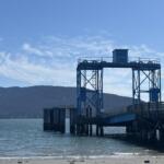Blue industrial pier structure stretching over the water with a mountainous coastline in the distance and a clear blue sky