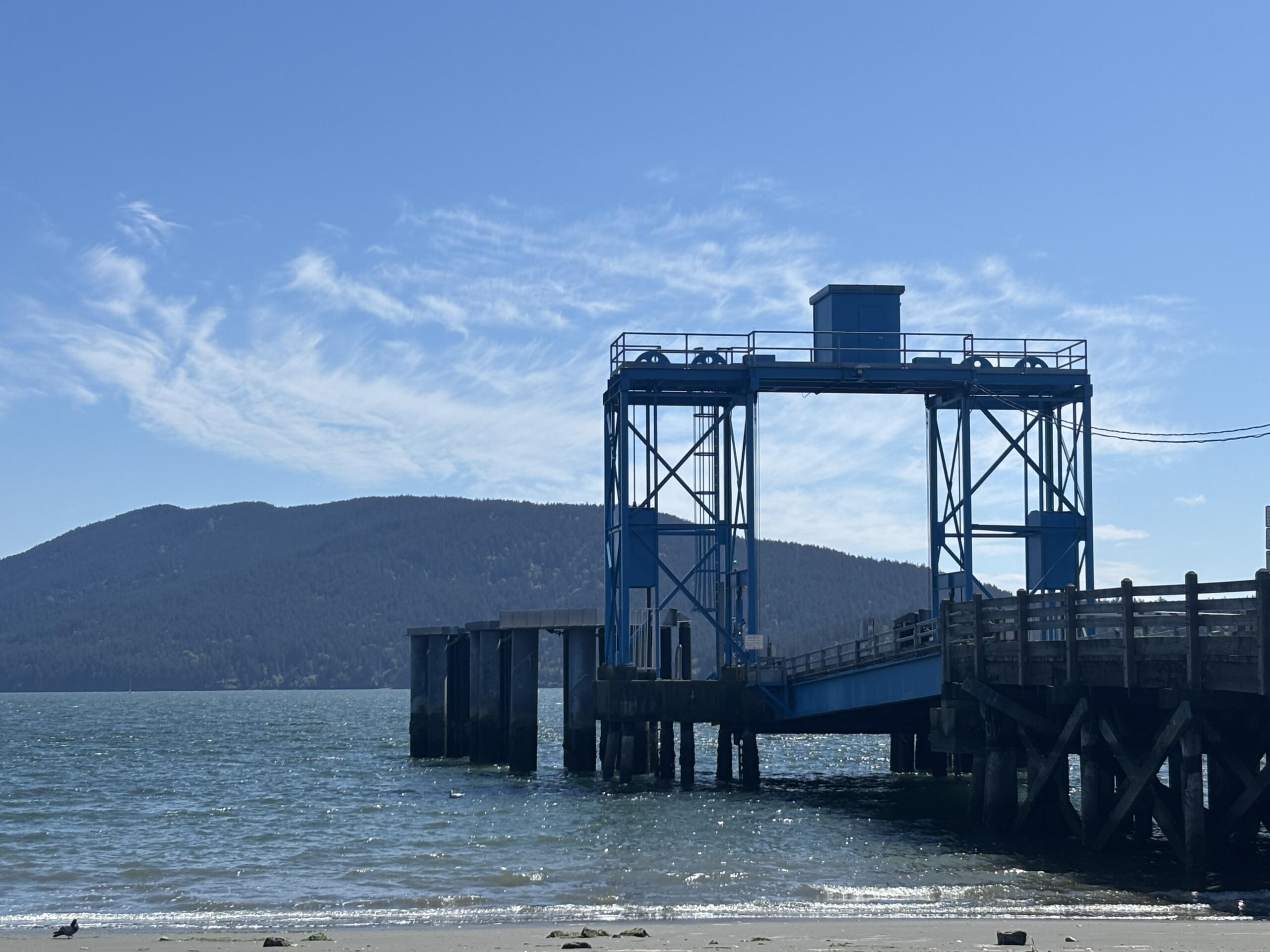 Blue industrial pier structure stretching over the water with a mountainous coastline in the distance and a clear blue sky