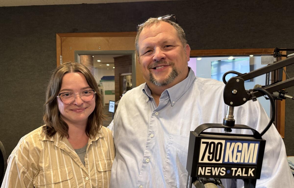 Two radio hosts pose in a studio, smiling at the camera with a large microphone in the foreground.