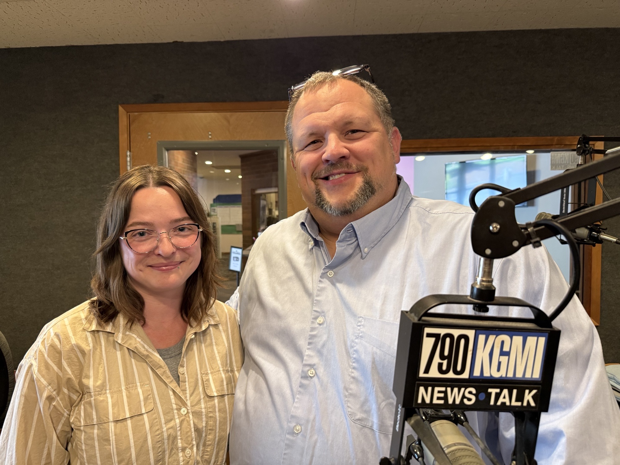 Two radio hosts pose in a studio, smiling at the camera with a large microphone in the foreground.